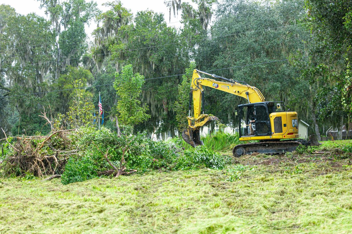 Excavator at the far edge of a freshly mowed field beside a pile of pushed-up brush
