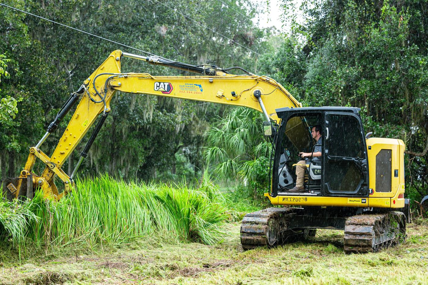 Excavator low in tall grass with its arm reaching to the left and the operator visible in the cab