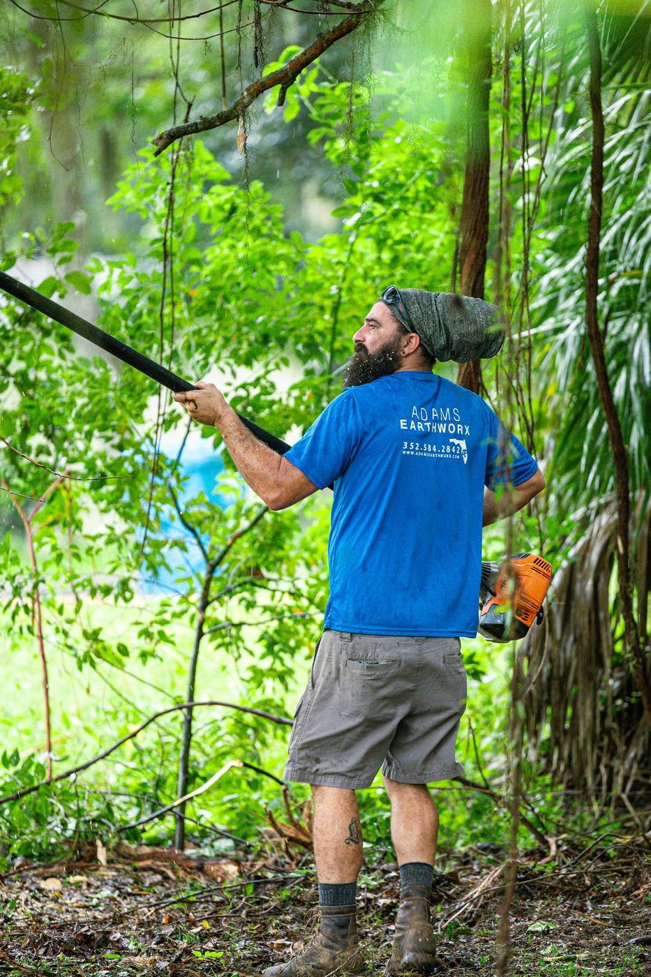 Crew member in an Adams Earthworx shirt holding a pole saw in dense green brush